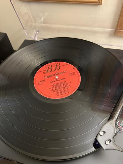 A close-up of a vinyl record playing on a turntable. The record features a red label with the title "Beggars Banquet" and the song "Face in the Crowd." The turntable shows part of the tonearm and cartridge.