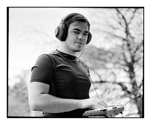 A black and white, square film portrait of a young man wearing headphones and holding a small computer keyboard outdoors.