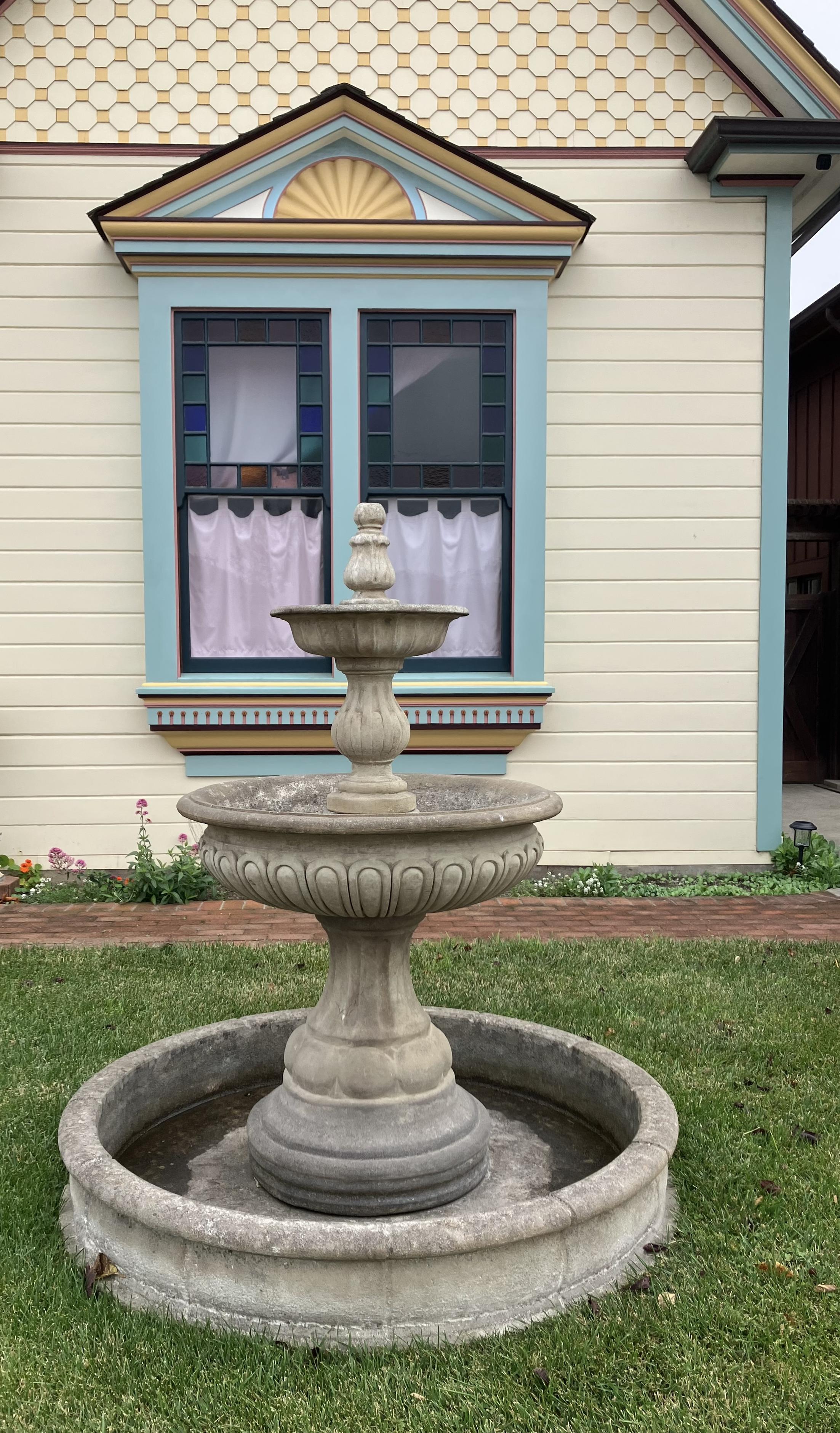A window which protrudes from the building, with four square panes. In the lower two some white curtains have been hung. The top two have lovely colored glass squares all around the edge of the window pane, leaving a small space in the center. Above the window is a triangular space with a sunburst inside it. There's a stone fountain in the yard in front of the window.