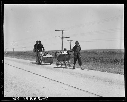 The black and white photograph depicts a group of people walking along an open road, pulling loaded vehicles behind them. Two men are leading the way; one man is pushing a cart filled with various items such as clothing and blankets while wearing a dark coat, hat, and gloves, suggesting it might be cold outside or that they may have traveled from another place where temperatures were lower than in Texas.
The second man is pulling what appears to be a baby stroller containing an infant. He wears a light-colored shirt under his jacket, darker pants, and shoes with laces; he also has on a dark hat.
Following them are two more individuals: one walking alongside the cart pushing it while wearing a coat and trousers in muted colors, likely indicating they have traveled significant distances or endured harsh conditions to reach this point. The other person is pulling the stroller carrying what seems like a child wrapped up warmly; both wear coats suitable for cool weather.
In the background are power lines extending into the distance on either side of an empty road that stretches far ahead. Sparse vegetation and open fields suggest a rural setting, possibly in Texas or another arid region with wide-open spaces typical of areas affected by droughts during periods like 1930's depression era depicted here.
The presence of these refugees indicates they might have been displaced due to economic hardships, likely as part of the large [...]