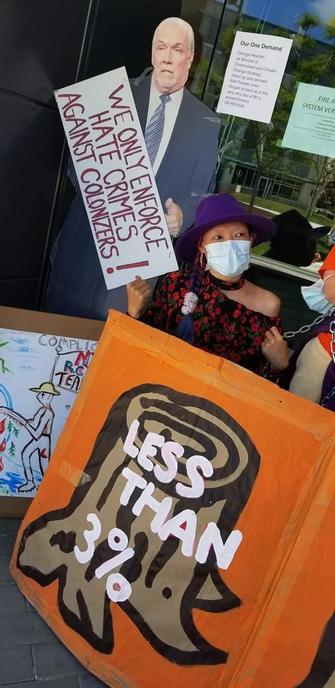 A person wearing a purple hat and a face mask holds an orange sign that reads, "LESS THAN 3%" against the backdrop of a building. Above them, a large cutout of a man in a suit holds a sign that reads, "WE ONLY ENFORCE HATE CRIMES AGAINST COLONIZERS!". A sign in the background states, "Our One Demand George Heyman as Minister of Environment and Climate Change Strategy stand up and demand that Premier John Horgan protect all of the very very last of BC's ancient forests OR RESIGN." The person is wearing a black floral print shirt. There are also two other signs in the background with text that can not be read in the image.