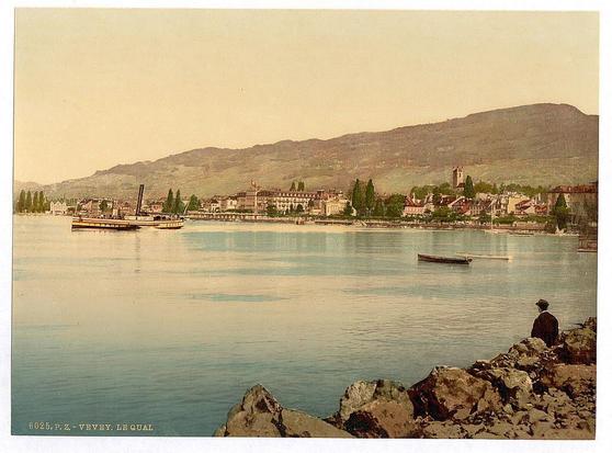The image depicts a scenic view over what appears to be Lake Geneva, in Vevey, Switzerland. In the foreground, calm waters reflect an old-fashioned steamboat and other boats moving across its surface. Onshore, buildings with European architecture line the lakeside, suggesting a quaint town setting with historical significance.

In the middle ground, there's another smaller boat on the lake, creating ripples as it travels along. The backdrop is dominated by rolling hills or low mountains under a slightly hazy sky, implying either early morning mist or light pollution in this serene environment.

On the right-hand side of the image stands an individual with their back to us; they are dressed in what looks like late 19th-century attire and wear a wide-brimmed hat. This person appears contemplative as if taking a moment to enjoy the vista before them, adding a human element that conveys leisure or vacation.

The overall color palette of this photograph is muted with sepia tones suggesting age; it evokes nostalgia for a bygone era in Switzerland's history.