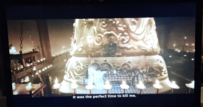 A couple of fighters protected by a spiritual force bell, in a room packed with candles. In the forground is a shelving unit with the shelves hung in chains from the ceiling instead of mounted on poles from the ground. All shelves have decorative carvings hanging from their undersides.