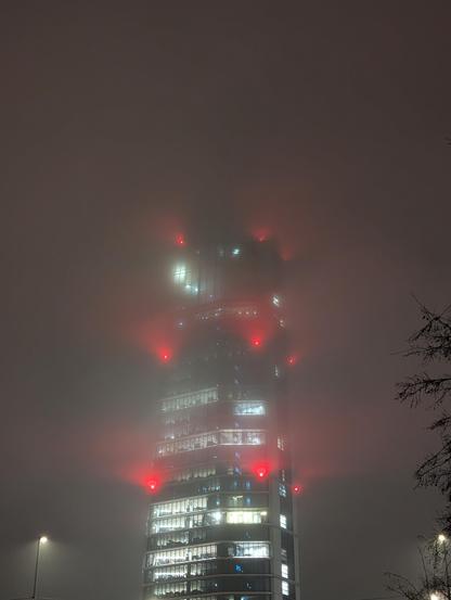 The MOL campus, a 28 storey tower, lit up during a foggy evening with multiple red lights placed evenly on the sides of the tower.
