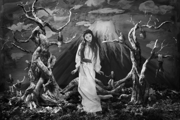 Photographie en n&b de Féebrile, mise en scène surréaliste avec le Mont Fuji peint en arrière-plan, ciel et nuages peints, et une femme au premier plan, avec un maquillage de type geisha, long cheveux noir et longue robe claire avec ceinture foncée, des arbres morts tous torturés de chaque côté et des tous petits humanoïdes un peu partout