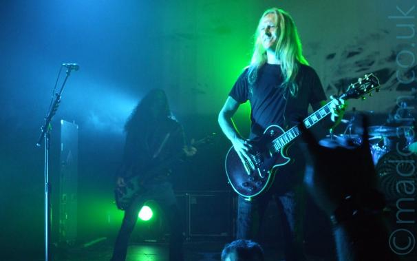 A man with long blonde hair, wearing a black t-shirt and jeans, and playing a black electric guitar standing on the right of the frame, with a microphone on a stand on the left.
In the background, there is a person with long dark hair , wearing a leather jacket, and playing a black and silver bass guitar, standing on the left of the frame, shrouded in a haze of smoke and green and blue lights.
A drum kit is visible in the background on the right, in front of a large backdrop where a video is being projected.
The whole frame has a blue and green cast from all the stage lights.