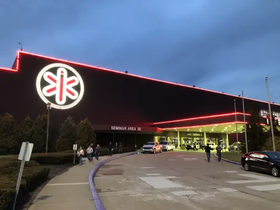 The image shows a building with a prominent sign featuring a stylized logo that has a lighted outline. The building is illuminated with red lights along its edges, and there are people walking in the vicinity. The entrance area appears to have a covered area where vehicles can pull up. It's likely a venue for events or seminars, as indicated by the signage mentioning a seminar area. The sky is overcast, suggesting that it is either dusk or early evening.

Image Credits: Wikimedia / Cards84664 / CC BY-SA 4.0
