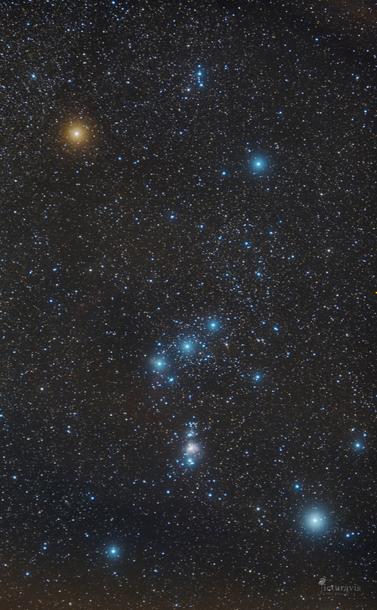 An image of the starry night sky focused on eight bright stars forming the Orion constellation. The top-left star is yellow, while the others are blue. Many more stars are visible, as well as the Orion Nebula, which is a small white blob in the lower center of the constellation.
