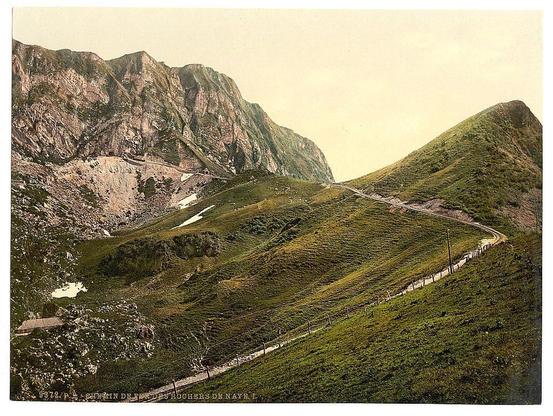 The image depicts a scenic mountainous landscape with lush green slopes and rocky outcrops. A winding path or road cuts through the terrain, suggesting accessibility to higher altitudes. The vegetation appears dense in some areas, indicative of varying altitude zones within the mountains. In the foreground, there are signs of human presence such as fencing alongside certain parts of the pathway. The sky is overcast with a soft glow that suggests either dawn or dusk lighting conditions. Notably absent from this image are any visible people or animals, which enhances its focus on natural beauty and solitude.