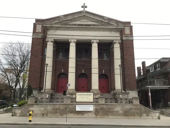 The image shows a building designed in a classic architectural style, likely a church, given the cross on top. It features a set of prominent columns and red double doors in the front, with a sign at the base that may indicate the name of the church and service times. The surrounding area includes some greenery and other buildings, with visible utility poles and wires.

Image Credits: Wikimedia / Sherlop / CC BY-SA 4.0