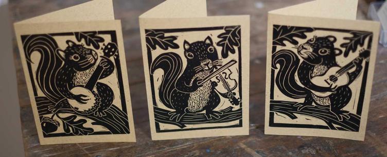 a photo of 3 hand printed linocut notecards depicting squirrels in an oak tree, playing a banjo, fiddle, and guitar