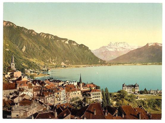 The image displays a picturesque European town, possibly located in the Alps region. The foreground shows densely packed buildings with red-tiled roofs and traditional architecture indicative of historical significance. Prominent among these structures is an old church or castle-like building featuring spires that pierce the skyline.

In contrast to this quaint human settlement lies Geneva Lake, its tranquil waters reflecting a light azure hue under what appears to be late afternoon sunlight. The lake's shoreline hosts larger buildings and perhaps residential areas with expansive grounds, one of which includes a large mansion-style estate overlooking the water.

The midground is dominated by steep green mountains blanketed in dense vegetation that gradually transitions into more rugged terrain towards the higher elevations where snow-capped peaks are visible against a clear sky. This suggests varying climate conditions within close proximity to each other and adds depth to the landscape depicted.

Overall, this scenic vista captures the harmonious blend of nature's grandeur with human habitation nestled in one of Europe’s most serene landscapes.