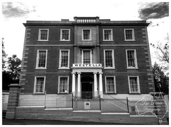 A powerful, straight on black and white photograph of Westella House, a three-story, symmetrical historic building with a central portico featuring columns.  The name 'WESTELLA' is visible above the main entrance. The foreground includes a low wall and black metal fencing.