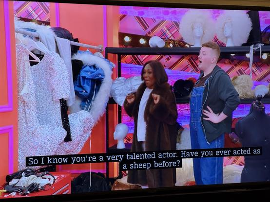 In the Drag Race UK werk room, Caitrin Feelings in boy mode on the right, with overalls and a darker top, says to Diane Parish on the left, in a (fake?) fur coat and a white top, "So I know you're a very talented actor. Have you ever acted as a sheep before?"