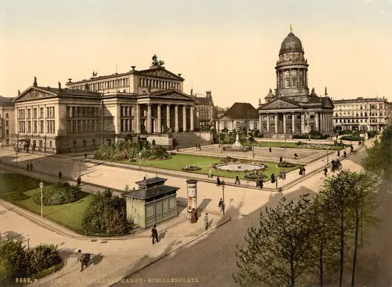 The image appears to be a historic, colored photograph of a public square, possibly featuring buildings of architectural significance. You can see a large building with steps, which might be a civic or cultural institution, alongside a notable round dome structure. There are also people walking around the square, and a landscaped area with flowers. The scene suggests a vibrant urban setting, likely from the late 19th or early 20th century.

Image Credits: Wikimedia / Unknown authorUnknown author / Public domain