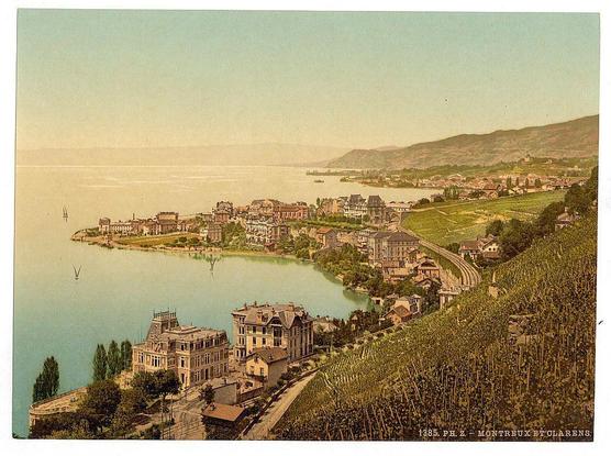 This image depicts a picturesque landscape overlooking Lake Geneva in Montreux, Switzerland. The photograph appears to be an antique postcard or print, given its vintage style and the presence of text indicating a location and years that suggest it's from around 1890-1900. It shows various buildings with distinct architectural features, such as mansions with intricate details near the lakefront and more modest structures further inland. The shoreline is lined with lush greenery and trees, while vineyards can be seen on elevated land in the foreground to the right.

The body of water reflects a light blue hue, indicating calm weather conditions at the time it was captured. Sailing boats are visible on the left side near the shore, adding life to the tranquil scene. The background reveals rolling hills or mountains that fade into a soft gradient towards the horizon under a sky with muted colors ranging from pale yellowish-green in the upper areas to lighter blue tones closer to where the lake meets the land.

The overall composition of this image suggests it was taken during the late 19th century, capturing a serene moment along Lake Geneva's shores. The presence of buildings and infrastructure implies that Montreux had developed into an established settlement by then.