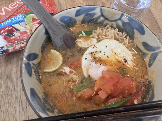 A bowl of flavorful ramen soup featuring a poached egg, lime slices, chopped tomatoes, and green chilli, with instant noodle packaging stating "Tom Yum Shrimp Flavour" in the background.