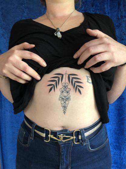 Photo du buste de Manon, sur le haut de son ventre est tatouée une fleurs de glycine, encadrée de deux feuilles