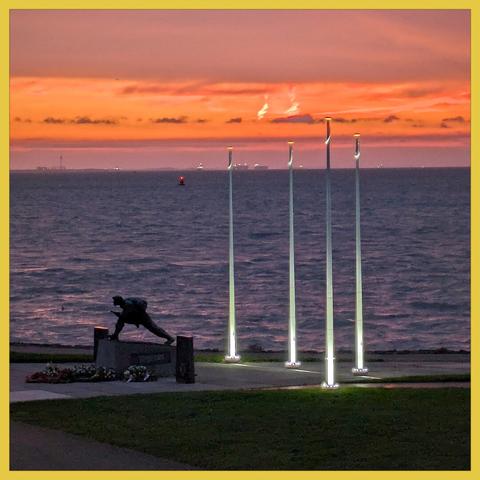 The monument of the liberation of Vlissingen (1944) stands in the early morninglight. The sky is very orange. Aside trhee illuminated flagpoles, without flags. In the background the water of the Western Scheldt