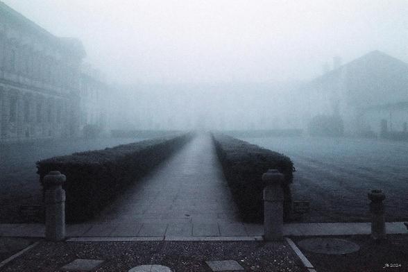 Blasses Farbfoto, das den Zugang zur Certosa di Pavia an einem nebligen Dezembermorgen zeigt. Der gepflasterte, links und rechts von einer Hecke gesäumte Weg ist noch am besten erkennbar, während die Gebäude links und rechts und im Hintergrund im Dunst kaum erkennbar sind. Leica R4 mit 35 mm Summicron auf Purple-Film.