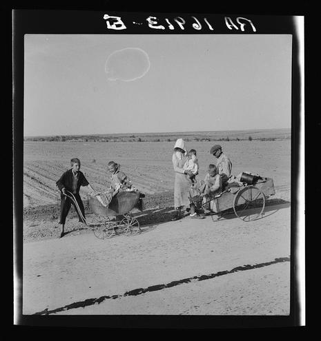 The photo shows a black and white scene with six individuals in an open field. Five of them appear to be part of a family, including three adults—a woman standing on the right wearing a sunhat, another seated on a homemade cart beside her holding two children who are sitting on what looks like straw bales or similar materials for cushioning—and a man at the far left operating a manual seed drill. The ground is cracked and dry with no visible vegetation apart from some distant trees in the background. In front of this family, there's another individual pushing a wheeled cart containing two more children who are looking directly towards the camera. Above them on what seems to be an airship or balloon, text reads "ELIPSI 17". The overall setting suggests rural work and perhaps migration during challenging times for farming communities in America during the Great Depression era.