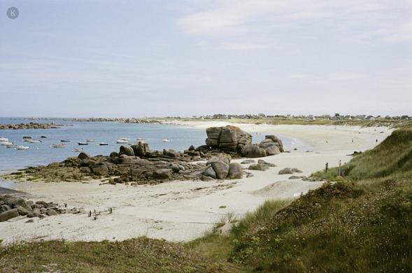 Strandlandschaft mit hellem Sand, verstreuten Felsen und türkisblauem Meer; kleine Boote liegen vor Anker, wenige Menschen spazieren am Ufer, dahinter sanfte Dünen unter leicht bewölktem Himmel.