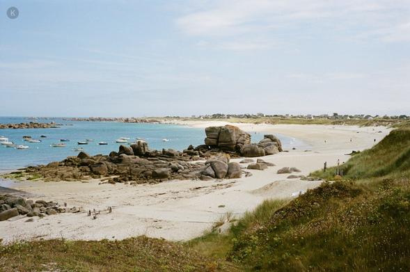 Strandlandschaft mit hellem Sand, verstreuten Felsen und türkisblauem Meer; kleine Boote liegen vor Anker, wenige Menschen spazieren am Ufer, dahinter sanfte Dünen unter leicht bewölktem Himmel.