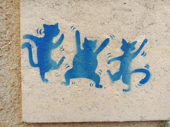 Street art qui représente 3 silhouettes de chat bleues en train de danser. Celui du milieu est de face, avec les 2 pattes avant levées. Les 2 autres chats sont de profil et tournés vers celui du milieu. Le street art est peint sur un mur en pierre beige/blanc cassé.