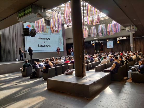 A packed room with people with a slide in front glbuenvenuti e buenvenute. Branded in Google colors.