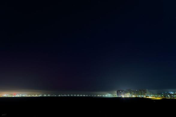 Das Foto zeigt eine nächtliche Szene mit einem weiten, dunklen Himmel, unter dem eine gerade Reihe von Straßenlaternen und beleuchteten Gebäuden am Horizont zu sehen sind. Auf der rechten Seite sind mehrere größere, beleuchtete Gebäude auszumachen, während links und mittig die Lichter in einer Linie angeordnet erscheinen. Der Himmel nimmt einen Großteil des Bildes ein, dominiert durch Dunkelheit und vereinzelt schwache Sterne, während die Häuser und Laternen mit ihrem Licht und Schein im aufziehenden Nebel einen klaren Kontrast bieten.