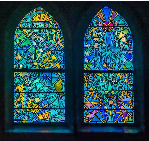 Two tall arched stained glass windows featuring abstract geometric designs in vibrant blues, teals, yellows, oranges, and purples. The left window contains flowing angular patterns with eye motifs scattered throughout. The right window displays a radial sunburst pattern at the top with a central cross motif, and geometric eye symbols in the lower sections. Both windows are divided into three horizontal sections by dark metal frames and are set against a dark wall, creating a luminous effect