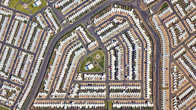 A satellite picture of a suburban neighborhood with uniformly designed white-roofed houses arranged in a grid-like pattern. Streets curve and intersect symmetrically. At the center, there is a green space featuring a circular structure, pathways, and a few buildings.