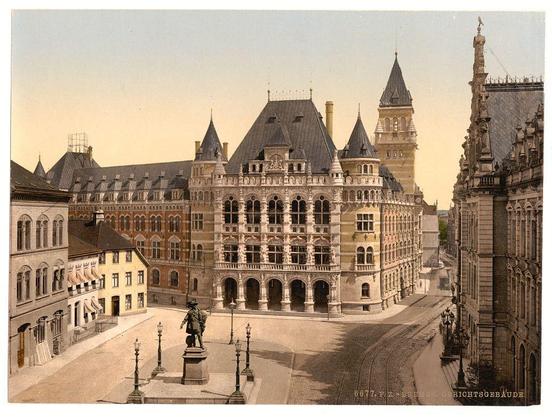 This image features a detailed view of the Palace of Justice in Bremen, Germany. The photograph captures an expansive courtyard with European architecture prominently displayed on multiple buildings surrounding it. These structures exhibit characteristic steep gabled roofs and ornate facades typical of 19th-century design.

A central focal point is a statue atop a pedestal, centrally located within the open area of the square. Surrounding this are several lampposts that add to the historical ambiance. The photograph has sepia-toned coloring indicative of its likely origin in the late 19th century or early 20th century.

The sky above is clear with no visible clouds, and there's a slight haze which may be atmospheric perspective or pollution from industrial era levels of pollutants. In the background are more buildings with windows reflecting sunlight, creating contrast between lit and shadowed areas on their facades. The overall composition conveys historical significance and urban planning characteristic of that period.

The image appears aged, as is evidenced by discoloration and wear marks visible in places suggesting its longevity and preservation from an era where such documentation was valued for capturing the essence of public architecture at a time when Bremen's growth had seen significant expansion.