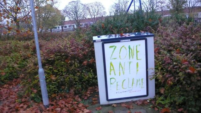 omringd door struiken, begroeiing, bomen in herfst kleuren oranje groen, een straat paal en een straakastje met reclamebord aangepast met witte poster met groene letters, zie caption