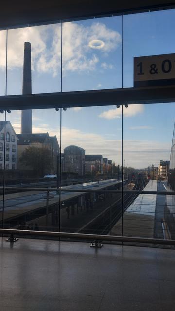 Looking through the glass wall of the station concourse, train tracks & platforms below, blue skies above, an old, tall, industrial brick chimney from a distillery on the left