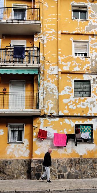 Una edificio con la fachada amarilla llena de mancha blancas. Al izquierda hay balcones con toldos verdes y a la derecha, ventanas. Un hombre camina por delante del edificio y en la planta baja hay ropa tendida.