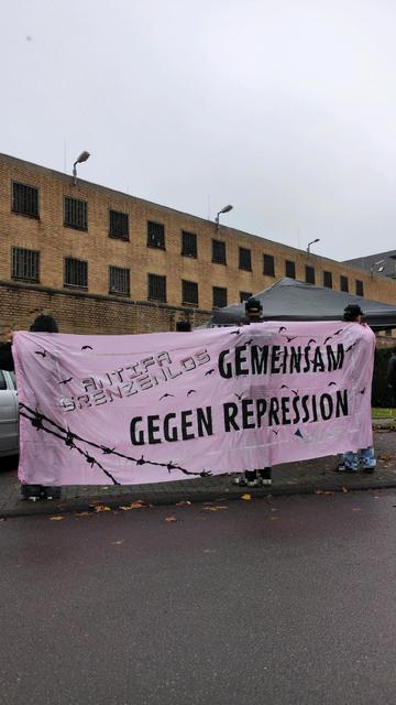 Menschen halten ein Transparent der Kampagne NIKA NRW mit der Aufschrift "Antifa Grenzenlos. Gemeinsam gegen Repression". Im Hintergrund ein Zellenblock der JVA Dinslaken.