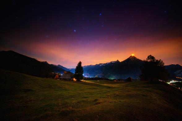Nachtaufnahme einer weiten, schwach beleuchteten Wiese im Vordergrund, die zu einem Tal und einer Bergkette hin abfällt. In der Mitte steht eine einzelne, dunkle Tanne, hinter der ein kleines, beleuchtetes Haus erkennbar ist. Im Hintergrund erheben sich hohe Berge, deren Silhouetten scharf gegen den leuchtenden Nachthimmel abzeichnen. Der Himmel zeigt einen tiefen Farbverlauf von dunklem Violett oben bis zu einem kräftigen Orange-Gelb über den Bergen, wobei die Sterne durchscheinen. Die Spitze des höchsten Berges auf der rechten Seite ist hell orange beleuchtet. Am rechten Bildrand sind die Lichter eines Dorfs zu sehen.
