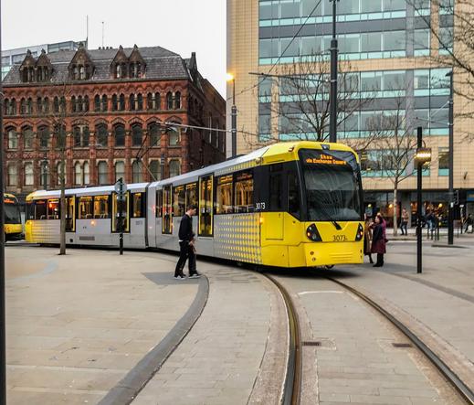 Eine Straßenbahn fährt auf einem Platz um eine Kurve. Sie ist grau/gelb und die Türen fangen erst in einer Höhe von etwa einem halben Meter überm Boden an