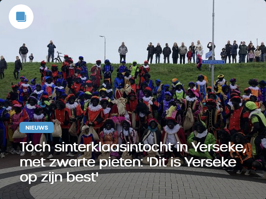 Een foto van een grote groep zwarte pieten op een pleintje voor een dijk met daarop een aantal toeschouwers.