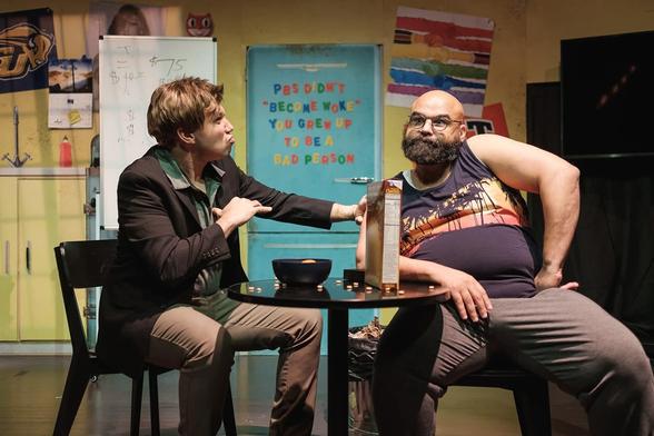 happywaterbear on stage as character Jake seated across from Stephen Emerson as the character Tim while having a comedic intense discussion about who's turn is it to take out the trash. Between them is a black table with bowl and box of cereal. In the background is a artificial set of the apartment. The refrigerator graphic has magnetic letters on the fridge saying "PBS didn't 'become woke', you just grew up to be a bad person" 

photo by caleb knight photography