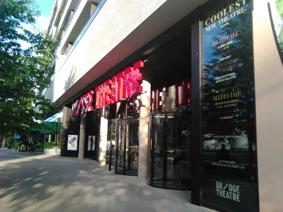The image shows the entrance of a theater building, which appears to have a modern architectural design. There are large glass doors leading into the theater, and prominent signage on the exterior that includes the words "BRIDGE THEATRE." Additionally, there are promotional posters or advertisements displayed on the side of the building, likely showcasing current or upcoming performances. The surrounding area has some greenery, indicating a pleasant outdoor environment.

Image Credits: Wikimedia / Koto / Public domain