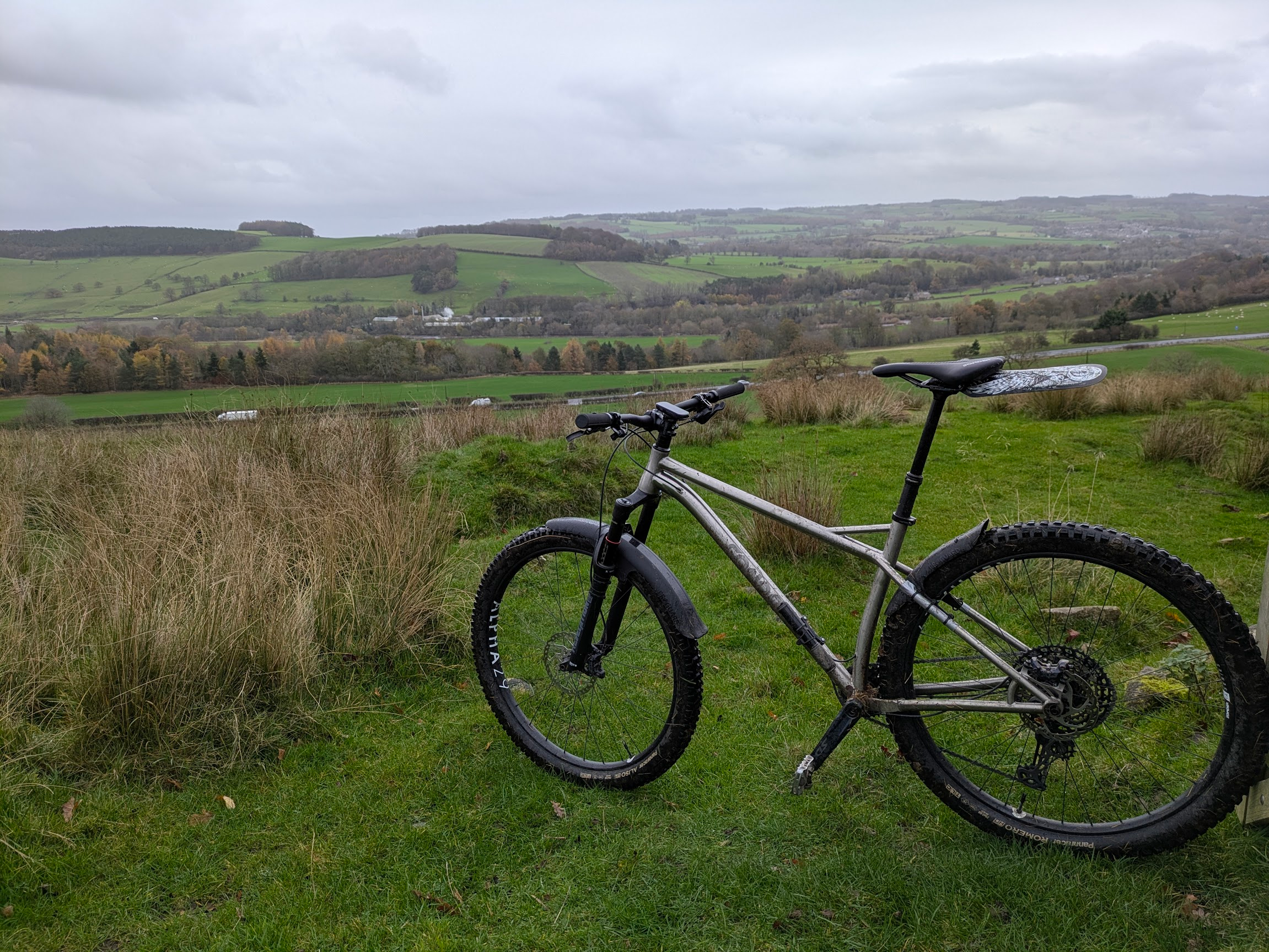Titanium mountain bike on a rough grassy field, with a view across a cloudy, grey Tyne Valley. A busy road runs along the bottom of the valley. In the distance, a small paper factory is making a cloud of steam.