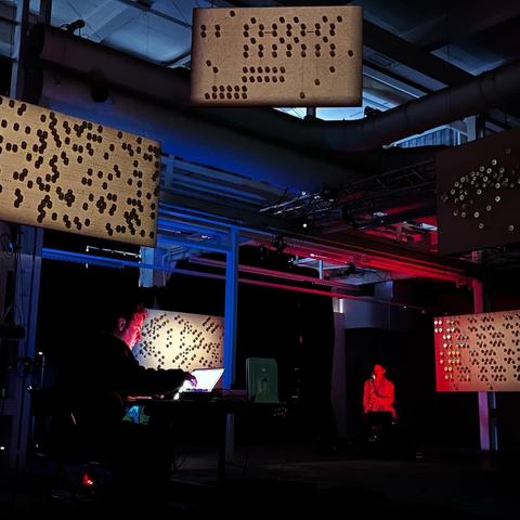 A darkened indoor space featuring a performer at a laptop and illuminated displays with abstract patterns. The setting includes colored lighting, creating a dramatic effect, while another seated figure is visible in the background.