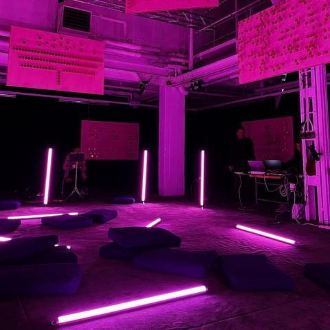 An art installation featuring vibrant pink lighting. The scene includes illuminated neon tubes, blue cushions scattered on the floor, and large pink panels hanging from the ceiling with various patterns. Two individuals can be seen in the background, one seated and the other at a