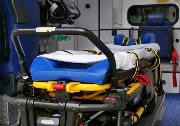 Trage mit blauem Polster und weißer Decke in einem Rettungswagen, medizinische Ausrüstung im Hintergrund.