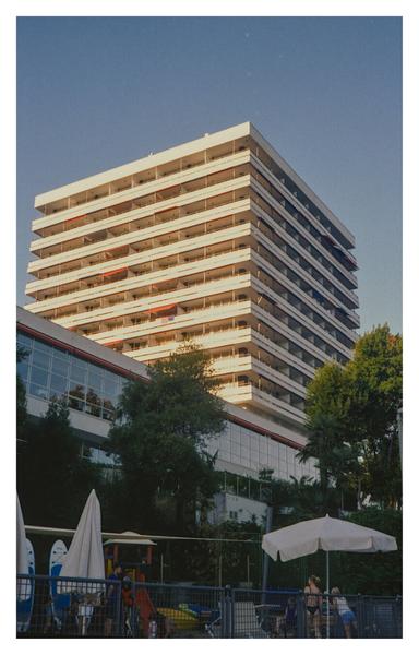 Color photograph of a modernist building on the coast of Opatija. The south side is already in shadow, while the west side is bathed in golden light and rises against the sky. In the foreground, in the shade, there is a small playground with a parasol and a few people.
