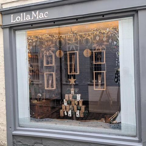 The LollaMac shop window decorated for Christmas. Grey wooden framed large shop window, with hanging wooden boxes displaying items of jewellery. In the centre of the window is a Christmas tree shaped display of items on small cards. The borders of the window are decorated with drawn-on festive foliage.