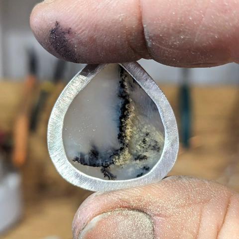 Rear view of a dendritic opal in an open-backed silver setting, held between grubby finger and thumb