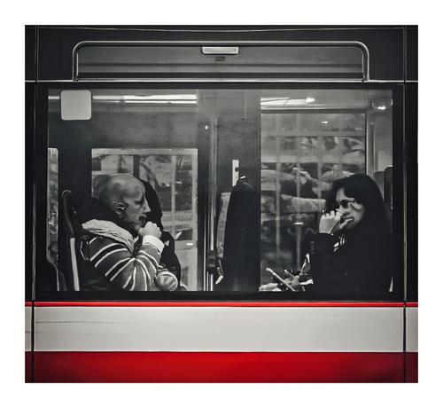 This is a photo of a bus interior, through a large window. A bald man with a warped face and striped jacket is sitting on the left. A woman with another warped face and long dark hair is wearing a black top while holding a phone, sits on the right. The bus window has reflections of the outside and interior lights. The bottom part shoes the red and white bus trim.